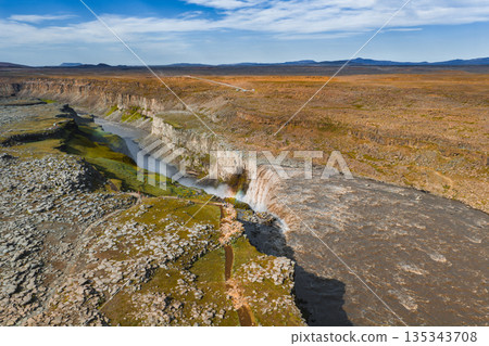Aerial view of Dettifoss waterfall in Iceland, with mist creating a faint rainbow. Rugged cliffs and barren highlands surround the powerful cascade. 135343708