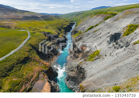 Studlagil Canyon in Iceland features a turquoise river, steep rocky cliffs, green vegetation, a dirt road, and a clear blue sky with distant hills. 135343710