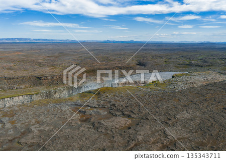 Aerial view of Dettifoss waterfall cutting through rugged volcanic terrain in Vatnajokull National Park, with misty spray and a winding path nearby. 135343711