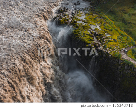 Dettifoss waterfall cascades forcefully into a deep canyon, with turbulent muddy water, moss covered terrain, and a visible path on the right. 135343718