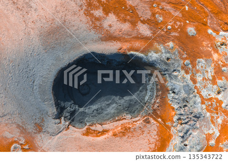 Aerial view of a dark mud pool surrounded by orange and reddish brown terrain, showcasing geothermal activity and textured mineral deposits in Iceland. 135343722