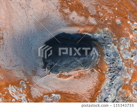 Aerial view of a bubbling geothermal mud pot in Iceland, surrounded by orange and gray mineral deposits. The terrain showcases vivid geothermal activity. 135343733