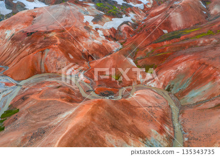 Vibrant rhyolite mountains in Landmannalaugar, Iceland, with red, orange, and brown hues, green moss, snow patches, and a winding river below. 135343735
