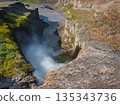Dettifoss waterfall cascades into a deep canyon with mist rising. Rugged cliffs, green moss, and a winding river define the dramatic Icelandic landscape. 135343736