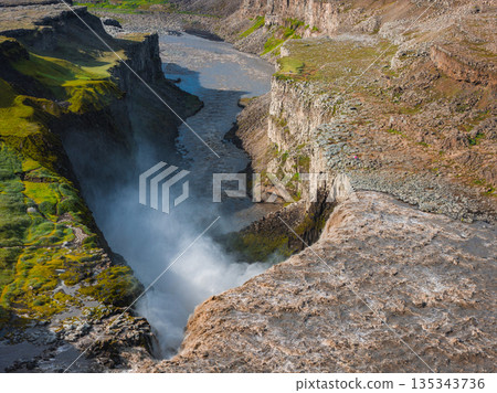 Dettifoss waterfall cascades into a deep canyon with mist rising. Rugged cliffs, green moss, and a winding river define the dramatic Icelandic landscape. 135343736