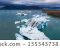 Aerial view of a glacial lagoon in Iceland with large icebergs in turquoise waters, rugged mountains in the background, and a cloudy sky overhead. 135343738