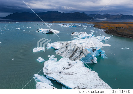 Aerial view of a glacial lagoon in Iceland with large icebergs in turquoise waters, rugged mountains in the background, and a cloudy sky overhead. Aerial view of a glacial lagoon in Iceland with large icebergs in turquoise waters, rugged mountains in the background, and a cloudy sky overhead. 135343738