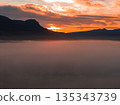 A vibrant sunset illuminates the sky with orange and red hues, silhouetting rugged mountains. Dense mist blankets the foreground, creating a serene scene. 135343739