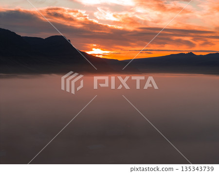 A vibrant sunset illuminates the sky with orange and red hues, silhouetting rugged mountains. Dense mist blankets the foreground, creating a serene scene. 135343739