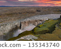 Dettifoss waterfall cascades into a misty canyon, surrounded by rocky terrain and moss, with a viewing platform under a vibrant sunset sky. 135343740