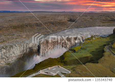 Dettifoss waterfall cascades into a misty canyon, surrounded by rocky terrain and moss, with a viewing platform under a vibrant sunset sky. 135343740
