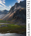 The Stokksnes Peninsula in Iceland features jagged mountains, green grasslands, reflective water channels, wetlands, and a small settlement below. 135343741