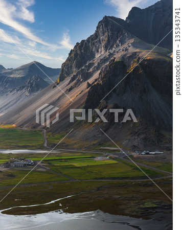 The Stokksnes Peninsula in Iceland features jagged mountains, green grasslands, reflective water channels, wetlands, and a small settlement below. 135343741