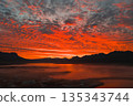 Fiery red and orange sunset over a calm body of water in Iceland, with silhouetted mountains, textured clouds, and a misty settlement in the foreground. 135343744