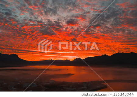 Fiery red and orange sunset over a calm body of water in Iceland, with silhouetted mountains, textured clouds, and a misty settlement in the foreground. 135343744