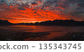 Fiery red and orange sunset over Icelandic mountains, mist covered ground, and a calm reflective body of water with a small settlement visible. 135343745