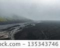 Aerial view of a volcanic landscape in Iceland with dark terrain, winding glacial rivers, and a moss covered mountain emerging from dense fog. 135343746