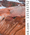 Aerial view of Iceland's Rainbow Mountains, featuring vibrant red, orange, and brown rhyolite formations with contrasting snow patches in the background. 135343752