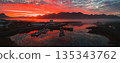 Aerial view of a small harbor in Iceland at sunset, with calm water reflecting red and orange skies, jagged mountains, mist, boats, and nearby buildings. 135343762