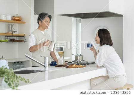 一對五十多歲夫婦的照片：一位男士在廚房煮咖啡，一位女士在喝咖啡。 135343895