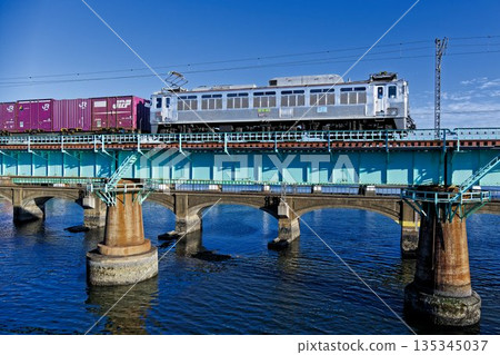 A freight train pulled by an EF81 electric locomotive crosses a railway bridge, its body shining in the setting sun. A freight train pulled by an EF81 electric locomotive crosses a railway bridge, its body shining in the setting sun. 135345037