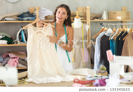 Smiling seamstress presenting handmade dress in sewing studio 135345633