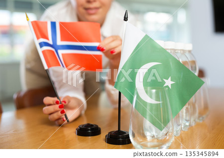 Young woman putting flags of Norway and Pakistan on table in office Young woman putting flags of Norway and Pakistan on table in office 135345894
