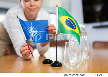 Woman secretary prepares an office for negotiations - she places flags of NATO (OTAN) and Brazil table Woman secretary prepares an office for negotiations - she places flags of NATO (OTAN) and Brazil table 135345939