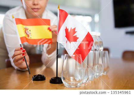 Young woman putting flags of Spain and Canada on table in office Young woman putting flags of Spain and Canada on table in office 135345961