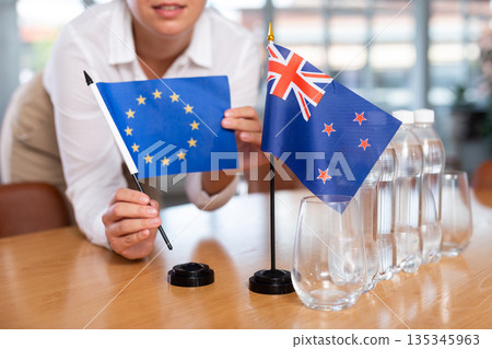 Young woman puts EU and New Zealand flags on table in office 135345963