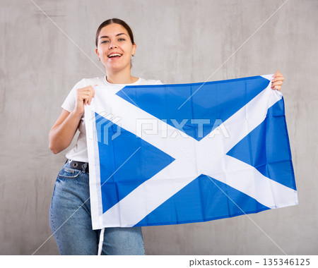 Joyous young woman holding Scotland flag against unicoloured background Joyous young woman holding Scotland flag against unicoloured background 135346125