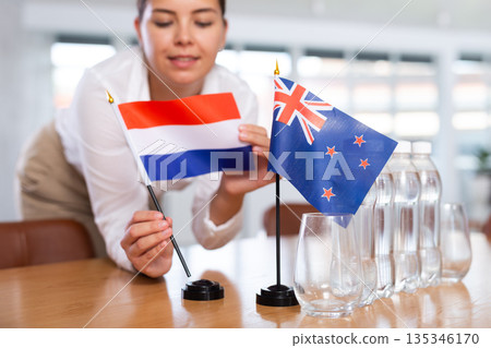 Young woman puts New Zealand and Netherlands flags on table in office 135346170