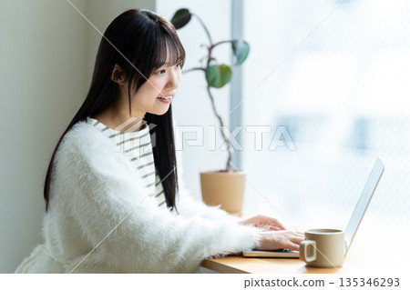 Smiling woman operating a laptop by the window Smiling woman operating a laptop by the window 135346293
