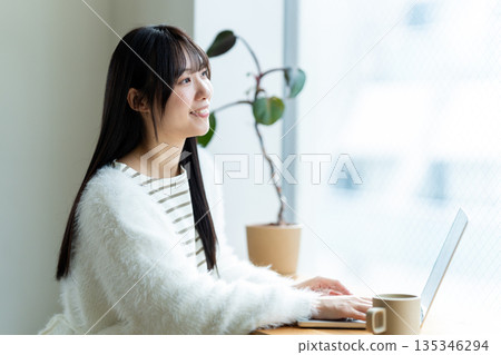 一位微笑的女子在窗邊使用筆記型電腦。 一位微笑的女子在窗邊使用筆記型電腦。 135346294