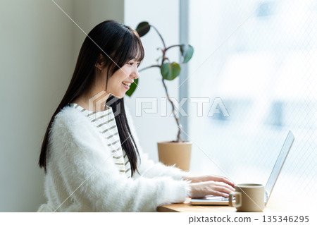 一位微笑的女子在窗邊使用筆記型電腦。 一位微笑的女子在窗邊使用筆記型電腦。 135346295