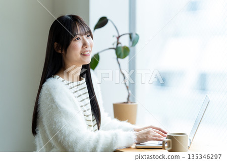 一位微笑的女子在窗邊使用筆記型電腦。 135346297