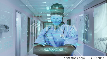 Standing doctor wearing light blue scrubs and mask in hospital hallway with holographic interface Standing doctor wearing light blue scrubs and mask in hospital hallway with holographic interface 135347094