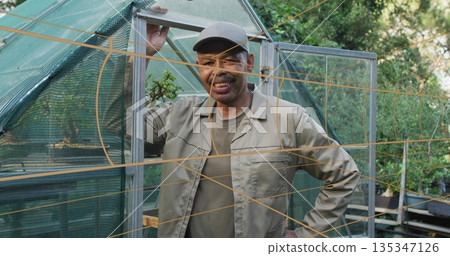 Mature gardener wearing gray cap and jacket holding greenhouse door in home garden with plant pots Mature gardener wearing gray cap and jacket holding greenhouse door in home garden with plant pots 135347126