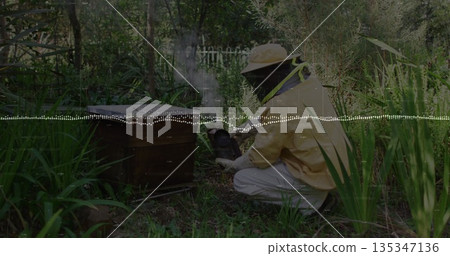 Crouching beekeeper wearing yellow suit using metal smoker beside hive box in garden, copy space Crouching beekeeper wearing yellow suit using metal smoker beside hive box in garden, copy space 135347136