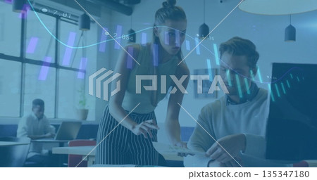 Teenage female wearing green top and colleague collaborating in open office, with printed charts 135347180