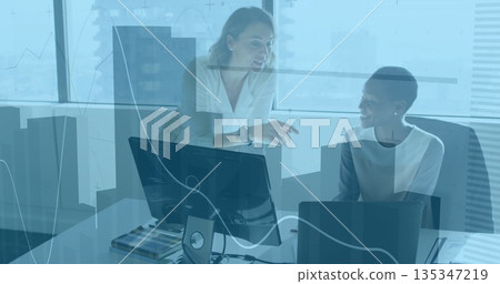 Collaborating women in business attire reviewing documents in office, with laptop, monitors Collaborating women in business attire reviewing documents in office, with laptop, monitors 135347219