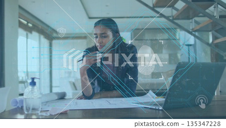 Leaning woman in blazer reviewing blueprints on desk in modern office, holding smartphone and pen 135347228