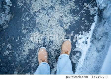 Person stands on asphalt sidewalk, winter road covered in mixture of technical salt and wet snow. 135347292