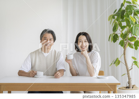 一對五十多歲的夫婦面帶微笑,並排坐在餐桌旁,看著鏡頭。 一對五十多歲的夫婦面帶微笑,並排坐在餐桌旁,看著鏡頭。 135347985