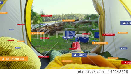 Relaxing camper lying in sleeping bag in yellow tent by lakeside clearing, with chairs and cooler 135348208