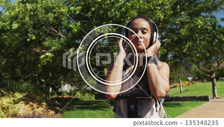 Balancing woman wearing tank top holding headphones to ears on scooter in park, with backpack 135348235