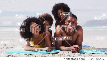 Lying family of four in swimwear posing on sandy beach towel, with social media icons overlay 135348237