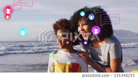 Standing mother in overalls and son in color-block shirt gazing at ocean shoreline, with chat icons Standing mother in overalls and son in color-block shirt gazing at ocean shoreline, with chat icons 135348255