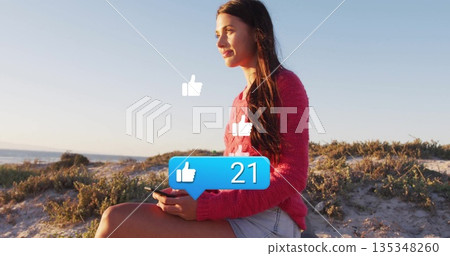 Sitting woman in sweater and shorts holding smartphone on dune viewing thumbs up icons, copy space 135348260