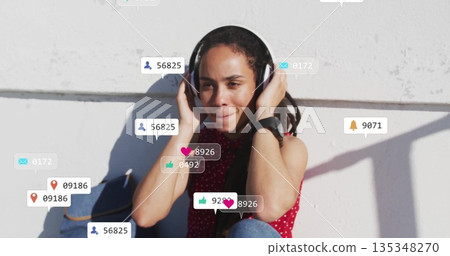 Sitting woman pressing headphones to ears on patio ledge, with social media notifications Sitting woman pressing headphones to ears on patio ledge, with social media notifications 135348270